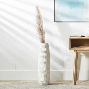 May include: A tall, textured white vase with a vertical, ridged pattern, holding a single, fluffy, beige pampas grass stem. The vase is placed on a grey rug. A wooden side table and artwork are in the background.