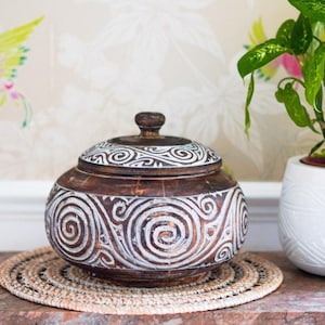 May include: A round, dark brown wooden lidded bowl featuring intricate, hand-carved white spiral and swirl patterns across its surface. The container has a small, rounded knob on top of the lid and a wide, bulbous body shape with a flat base.