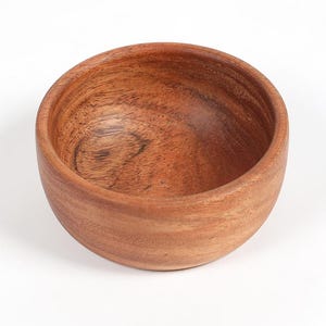 May include: A small, round wooden bowl with a natural wood grain finish. The bowl is empty and has a smooth, curved interior. The exterior is a warm brown color, and the bowl appears to be made of a single piece of wood.