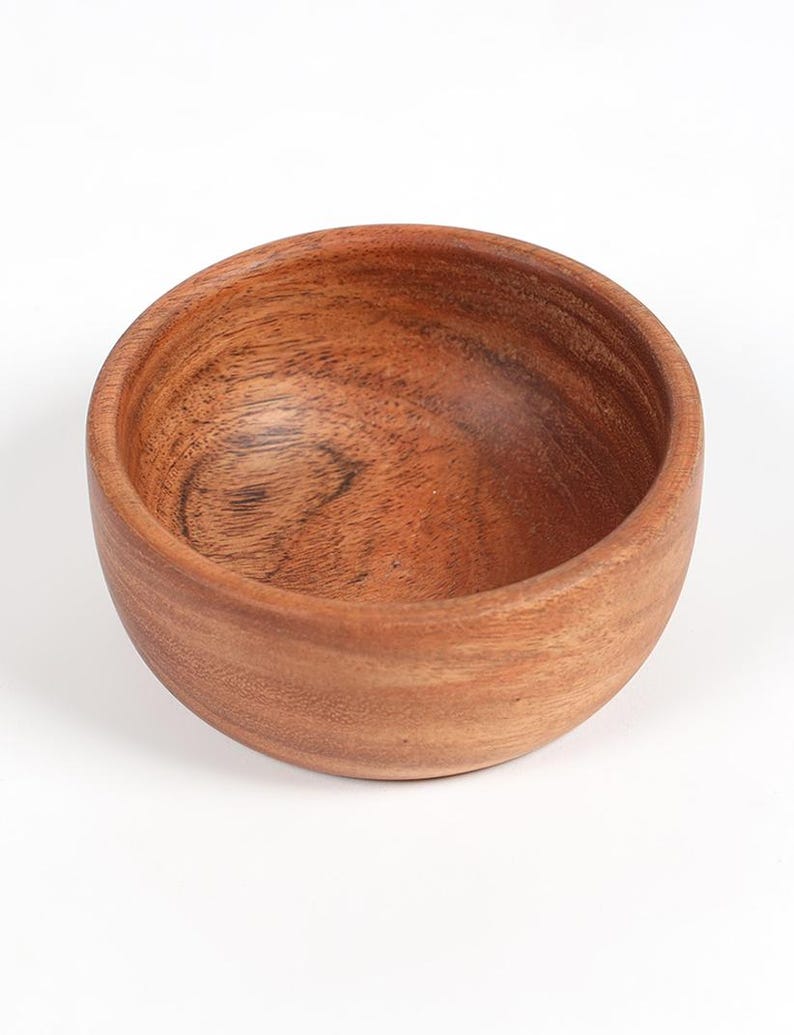 May include: A small, round wooden bowl with a natural wood grain finish. The bowl is empty and has a smooth, curved interior. The exterior is a warm brown color, and the bowl appears to be made of a single piece of wood.