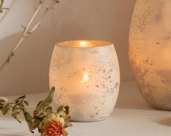 May include: A small, round, off-white candle holder with a textured surface and a gold interior. A lit candle glows inside. A second, larger candle holder is visible in the background. Dried flowers are in the foreground.
