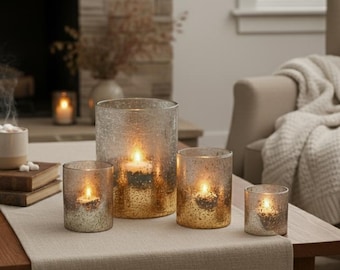 May include: A collection of four glass candle holders with lit candles inside. The holders vary in size and have a textured, crackled appearance. The glass has a gold and silver metallic finish, reflecting the warm glow of the candles. The arrangement sits on a neutral-colored table runner.