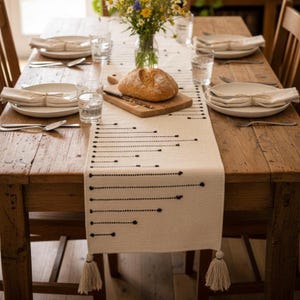 May include: A cream-coloured woven table runner featuring a black embroidered pattern of horizontal lines and dots, finished with tassels at both ends. The runner is placed on a wooden table with white plates, napkins, and a loaf of bread.