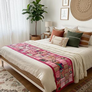 May include: A bedroom scene with a bed featuring a pink patchwork runner. The bed is dressed with pillows in brown, green, and cream tones. A woven rug and a large potted plant enhance the room's aesthetic.