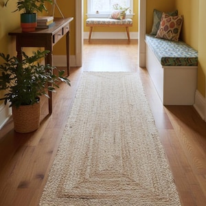 May include: A rectangular runner rug made of natural, light beige woven fibres featuring a subtle, concentric rectangular border pattern. The rug is positioned centrally on a polished wooden floor in a hallway, extending from the foreground.