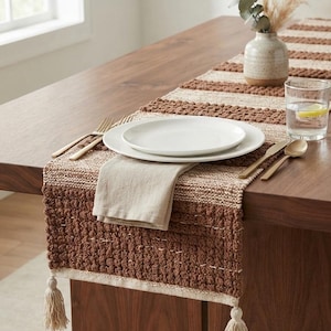 May include: A woven table runner featuring alternating horizontal bands of textured brown tufted fabric and smooth cream-coloured weave. The runner is finished with cream-coloured tassels at each corner and is displayed on a wooden table.