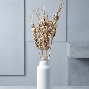 May include: A matte white cylindrical vase containing several stalks of metallic gold wheat. The wheat stalks feature detailed, pointed grains and thin, wispy tops. A small white tag with the text VAYU is attached to the stems.