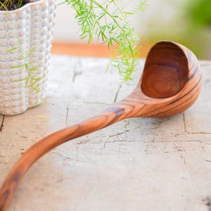 May include: A handcrafted wooden ladle with a long, curved handle and a deep bowl. The ladle is made of a rich, warm-toned wood, showcasing the natural grain. The kitchen utensil is designed for serving soups and stews.