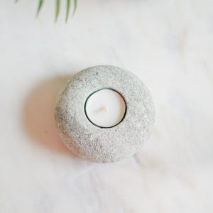 May include: A grey stone candle holder with a white tea light candle. The candle holder is round with a textured surface. The background is a white marble surface.