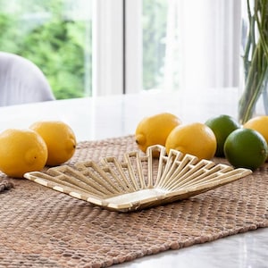 May include: A gold-coloured decorative tray with a fan-like design, placed on a woven runner. Fresh lemons and limes are arranged around the tray. The scene is set on a marble surface near a window.