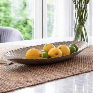 May include: An oval silver-coloured decorative bowl filled with lemons and limes. The bowl has a textured surface and a scalloped edge. The fruit is arranged on a woven table runner, with a vase of flowers in the background.