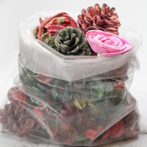 May include: A white mesh bag filled with dried potpourri. The bag contains a variety of decorative elements, including a pink rose, green pinecone, and other dried flowers and spices. The potpourri is a mix of red, green, and brown.