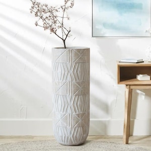 May include: A tall, light-coloured cylindrical vase with a geometric pattern. The vase holds a small tree branch with delicate pink flowers. The vase is placed on a rug in front of a white wall.