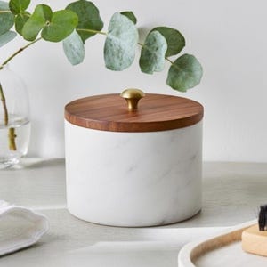 May include: A white marble storage jar with a wooden lid and a gold knob. The cylindrical container is on a light surface. Eucalyptus leaves are in a glass vase in the background.