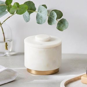 May include: A cylindrical white marble container with a matching lid and a circular knob handle. The base of the container is accented with a smooth, metallic gold band. The object features natural stone veining throughout the white marble surface.