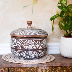 May include: A round, dark brown wooden lidded container featuring a white, hand-carved pattern of swirling spirals and geometric lines. The lid has a small, matching brown knob handle at the top, and the overall finish shows a distressed texture.