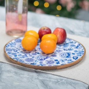 May include: A round wooden serving tray with a blue and white floral design holds oranges and red apples. The tray has a light brown wooden rim. A pink glass is in the background.