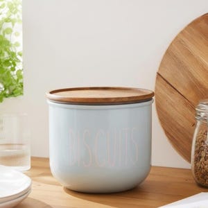 May include: A light blue cylindrical ceramic canister with a smooth finish and a natural wood lid. The word BISCUITS is printed in a thin, light-coloured sans-serif font on the front of the container, which has a rounded base and straight sides.