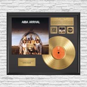 Op de afbeelding: Een ingelijste ABBA Arrival gouden plaat display met de albumhoes van de band in een helikopter, een gouden vinylplaat met een oranje label in het midden, een QR-code, een kleine albumafbeelding, een logo en een gepersonaliseerd naamplaatje.