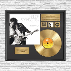Może przedstawiać: Oprawiona ekspozycja zawierająca okładkę albumu Born to Run Bruce'a Springsteena, złotą płytę oraz złotą tabliczkę z napisem YOUR NAME. Zestaw zawiera kod QR, małe zdjęcie oraz blok tekstu opisujący ten konkretny album muzyczny.