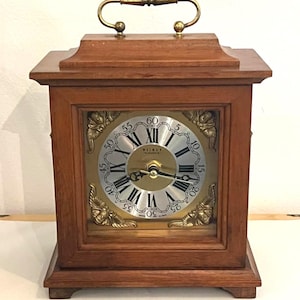 May include: A wooden mantel clock with a rectangular frame and a brass handle on top. The clock face features a gold centre, silver chapter ring with black Roman numerals, and gold-toned corner accents with cherub motifs. The text Weimar is visible.