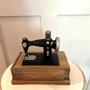 May include: A black sewing machine model with gold-coloured decorative scrollwork, a silver handwheel, and a blue thread spool on top. The machine is mounted on a rectangular wooden box base with a natural finish and visible wood grain texture.