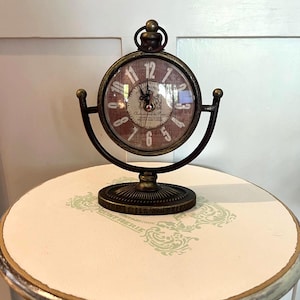 May include: A round tabletop clock with a dark bronze finish, featuring a distressed red face with white Arabic numerals and the text "Chateauneuf du Pape". The clock is suspended in a U-shaped metal frame mounted on a circular pedestal base.