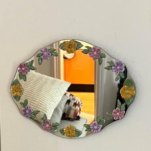 May include: An oval-shaped wall mirror with a scalloped edge, featuring a decorative border of sculpted, hand-painted ceramic flowers in shades of yellow, purple, and pink, interspersed with green leaves attached directly to the glass surface.