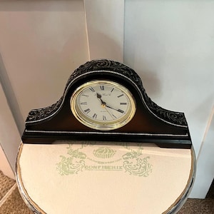 May include: A black mantel clock with a curved top and intricate carved scrollwork details. The clock face is white with black Roman numerals, gold-coloured hands, and a gold-toned circular frame. The base features a thin, dotted silver-coloured trim.