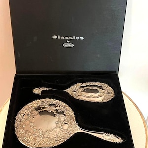 May include: A silver-toned vanity set featuring a round hand mirror and a smaller hairbrush, both with ornate embossed floral detailing. The items are presented in a black box with the text Classics By ILSAMUEL printed in silver on the lid.