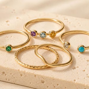 Op de afbeelding: Een verzameling van vijf goudkleurige ringen. Eén ring heeft een enkele groene steen, een andere heeft drie stenen in paars, blauw en geel, en een derde is een open ontwerp met een heldere steen en een turquoise steen. Twee effen ringen zijn inbegrepen.