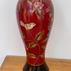 May include: A tall, slender vase with a deep red body and a black, speckled base. The surface features hand-painted illustrations of a small cream-coloured bird perched on a vine, accompanied by pink floral blossoms, green leaves, and red berries.