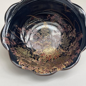 May include: A black bowl with a scalloped rim featuring an intricate gold and reddish-brown floral design painted on the interior. The artwork depicts detailed chrysanthemum flowers and foliage, creating a layered botanical pattern across the surface.
