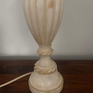 Peut inclure: Un pied de lampe de table en pierre blanc cassé avec des veines naturelles de couleur beige. La lampe présente un corps en forme d'urne, haut et cannelé verticalement, un piédestal central avec un détail sculpté en corde torsadée.