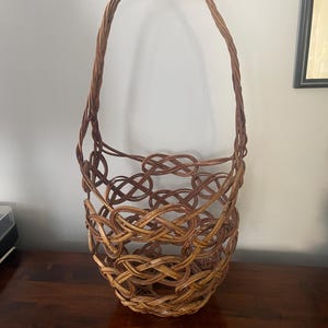 May include: A tall, open-weave basket crafted from brown, flexible willow branches. The structure features a series of interconnected, knot-like loops forming a cylindrical body, topped with a single, high, arched handle made of twisted wood.