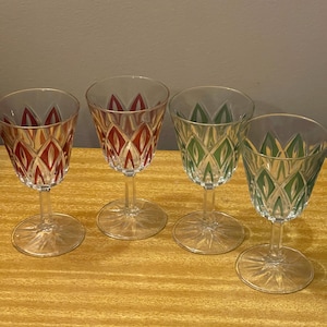May include: Four clear glass stemmed goblets arranged in a row. Two glasses feature a red diamond-shaped pattern, while the other two feature a green diamond-shaped pattern. Each glass has a clear stem and a circular base with a starburst design.