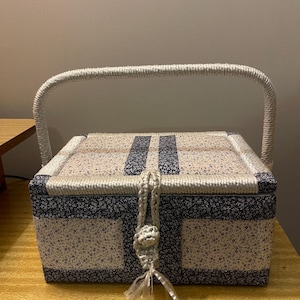 Op de afbeelding: Een rechthoekige naaidoos met een stoffen buitenkant in een crème en marineblauw bloemenpatroon. Het deksel wordt vastgezet met een gevlochten koordlus en een decoratieve sluiting. Een dik, gevlochten crème-kleurig handvat boogt over de bovenkant.