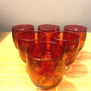 May include: A set of six ruby red glass tumblers arranged in a triangular formation. Each glass features a rounded body and a clear base, decorated with etched starburst patterns scattered across the exterior surface of the vibrant red glass.