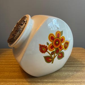 May include: A white ceramic canister with a slanted top opening and a textured cork stopper. The side of the canister features a colourful floral graphic with red, orange, and yellow petals, along with green leaves, arranged in a stylised botanical pattern.