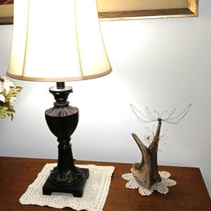 Könnte beinhalten: Eine dunkelbraune Tischlampe mit cremefarbenem Lampenschirm. Die Lampe steht auf einer weißen gehäkelten Tischdecke. Eine dekorative Drahtskulptur einer Blume befindet sich auf einem Holzsockel.