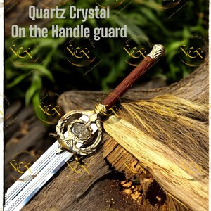 May include: A decorative sword with a quartz crystal on the handle guard. The sword features a gold-colored handle guard and a brown leather-wrapped handle. The blade is silver and the text "Quartz Crystal On the Handle guard" is visible.