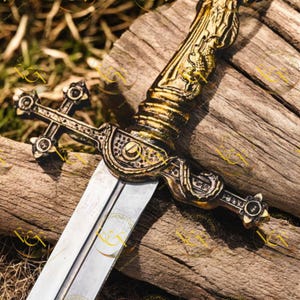 May include: A decorative sword with a silver blade and an ornate gold-colored hilt. The hilt features intricate designs and a cross-shaped guard. The sword rests on a weathered wooden surface, showcasing its detailed craftsmanship.