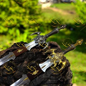 May include: Two fantasy swords with ornate silver and gold hilts and blades. The swords have intricate designs and are displayed on a dark, textured surface against a blurred green background. The blades have inscriptions.