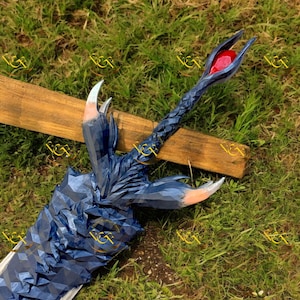 Puede incluir: Una espada de fantasía azul detallada con un diseño geométrico texturizado. La empuñadura presenta una gema roja y detalles en forma de garra. La hoja es plateada y la espada descansa sobre una tabla de madera.