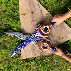 Puede incluir: Dos espadas de fantasía con un diseño de hoja azul y plateado y detalles dorados. Los mangos son azules con detalles circulares dorados. Las espadas se exhiben sobre una superficie de madera sobre hierba verde.