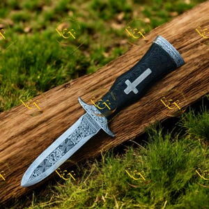 May include: A decorative dagger with a silver blade and ornate handle. The handle is black with a silver cross design. The blade features detailed engravings. The dagger rests on a wooden surface against a backdrop of green grass.