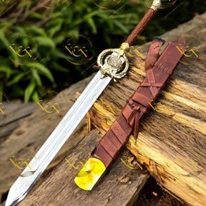May include: A decorative sword with a silver blade and a gold-colored hilt. The handle is wrapped in brown leather. The sword rests beside a brown leather sheath with a yellow tip. The image includes the text "Ebon Griffon Forge".