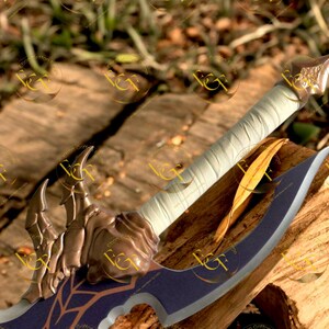 May include: A bronze and navy blue fantasy sword with a curved blade and ornate hilt. The handle is wrapped in white fabric. The sword rests on a wooden surface, showcasing its detailed design.