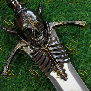 Może przedstawiać: Szczegółowe zbliżenie dekoracyjnego miecza z głowicą w kształcie czaszki i wzorem klatki piersiowej na rękojeści. Ostrze jest srebrne, a głowica i rękojeść są ciemnoszare. Miecz jest umieszczony na zielonym tle.