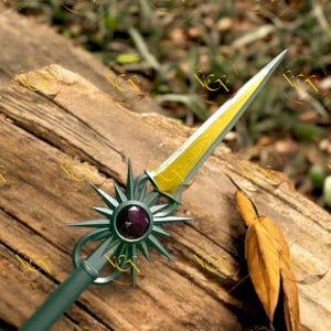 May include: A decorative spear with a silver blade and a gold-colored center. The handle is green with a starburst design and a purple gem. The spear is resting on a wooden surface.